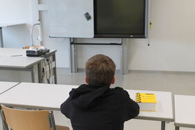 Junge sitzt an einem Schreibpult in der Schule. Einen Duden hat er neben sich auf dem Tisch liegen.