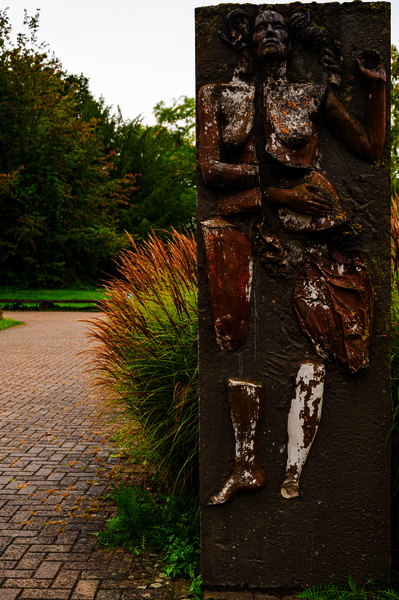 Das Bild zeigt eine Skulptur im Grünen. Die Skulptur selbst ist eine zerbrochener Körper der zu Teilen aus einer flachen Oberfläche hinausragt. 
