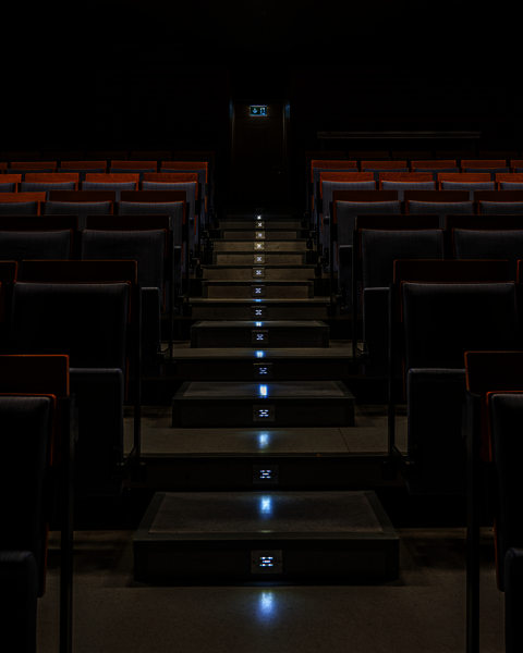 Eine Treppe in einem Hörsaal. Das Licht erhellt nur eine Reihe von Stühlen, während in den Stufen die Notbeleuchtung glimmt.