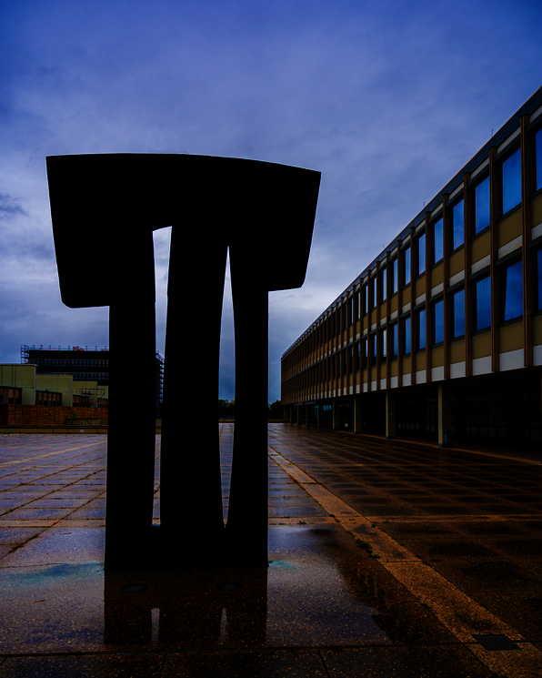Eine große Bronze-Skulptur mit einem Hammerartigen Kopf und zwei Aussparungen im Körper. Auf der rechten Bildseite ist die Fassade des Gebäude 1 der PH zu sehen.